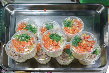Ready-to-eat Thai mixed vegetable salad (corn, crab stick, tuna) in plastic boxes on a metal tray. A convenient and healthy street food snack option.の写真素材