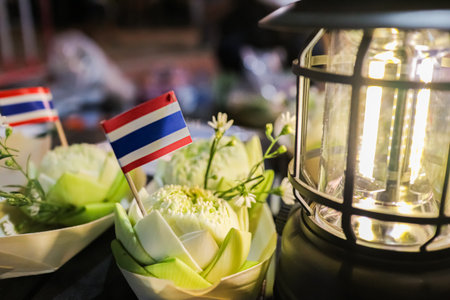 White lotus Krathong with a Thai flag placed near a glowing lantern, representing the calm night atmosphere of Thailandâs Loy Krathong festival and traditional rituals.の写真素材