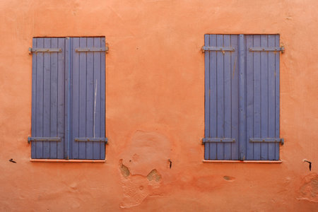 Bright orange wall features two worn blue shutters, adding a rustic touch to the architecture. The warm color evokes a serene atmosphere in a sunny environment.の写真素材