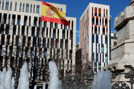 A vibrant flag flutters proudly in the wind amidst contemporary architecture in Madrid. A sculpted fountain with flowing water adds to the urban charm under a bright blue sky.の写真素材