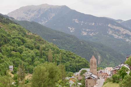 A quaint village nestled in the mountains features a historic church, lush greenery, and towering peaks under a cloudy sky. Nature and architecture harmonize beautifully in this serene landscape.の写真素材
