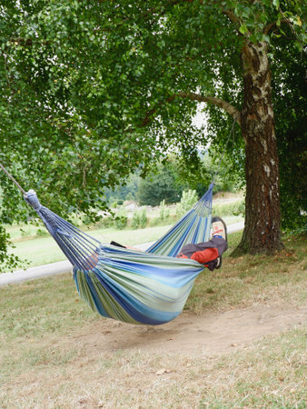 A person enjoys leisure time resting in a colorful hammock strung between two trees, surrounded by nature on a calm afternoon. The lush greenery adds to the peaceful atmosphere.の写真素材