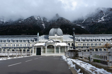 A grand, historic structure of train station stands amidst snow-covered ground and towering mountains. Low clouds loom overhead, adding to the serene winter atmosphere.の写真素材
