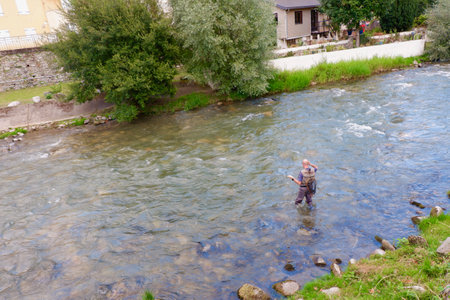 Unrecognised man stands in shallow water, casting his fishing line in a river as a serene rural landscape surrounds him under partly cloudy skies.の写真素材