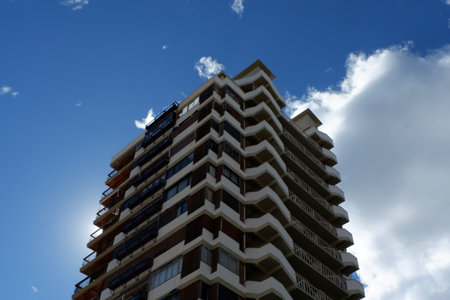 A tall residential building stands prominently against a vibrant blue sky, showcasing its contemporary design with distinctive balconies and sharp lines. The sunlight peeks from behind the structure, creating an eye-catching silhouette. Fluffy clouds drift across the sky, adding depth to the urban landscape.の写真素材