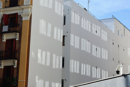 Morning sunlight casts intricate shadows of window frames on a white building in a lively neighborhood, highlighting the urban design and architecture.の写真素材