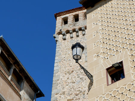 A tall stone tower rises against a clear blue sky, surrounded by charming buildings showcasing textured walls and a vintage lantern hanging from one of them.の写真素材