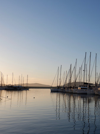 Sailboats docked in a marina capture the last light of sunset, their reflections shimmering on the calm water. A tranquil atmosphere surrounds the scene as evening approaches.の写真素材