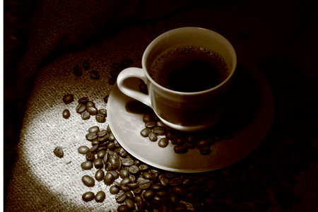 A cup of coffee standing on a burlap. Around scattered coffee beans In semi-dark lighting and balance in sepiaの写真素材