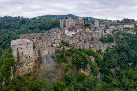 Aerial view of Sorano, Italy, showcasing medieval architecture nestled in lush Tuscan hills. Stone structures and fortified walls dominate this historic hilltop village under a cloudy sky.の写真素材