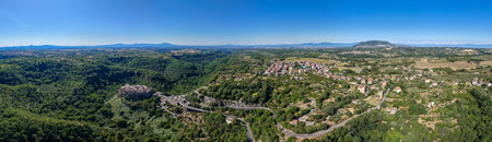 Aerial view of the medieval village of Calcata, Italy, perched on a cliff surrounded by dense forest under clear skies.の写真素材