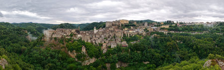 Aerial view of Sorano, Italy, showcasing medieval architecture nestled in lush Tuscan hills. Stone structures and fortified walls dominate this historic hilltop village under a cloudy sky.の写真素材