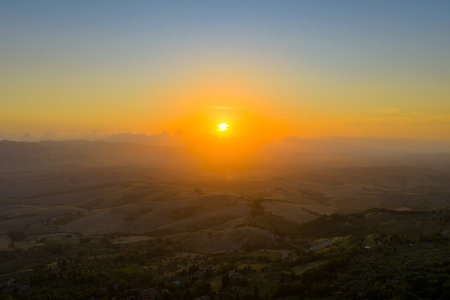 Aerial view of rolling Tuscan countryside near medieval Volterra at sunset, warm summer haze softening distant hills and fields.の写真素材