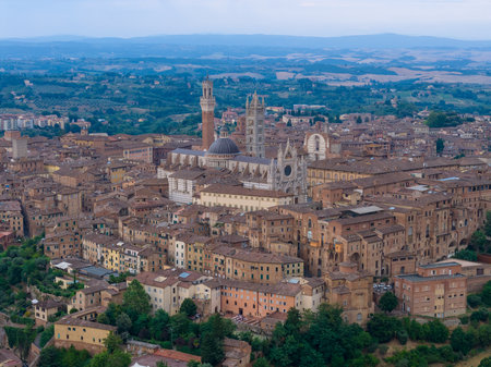 Aerial view of Siena, Italy, highlighting its medieval architecture and historic cityscape. Terracotta rooftops and the iconic Torre del Mangia stand out against the lushの写真素材