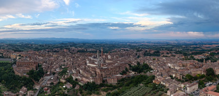 Aerial view of Siena, Italy, showcasing its medieval character with terracotta rooftops. Highlights include the Siena Cathedral and Torre del Mangia against a backdrop of Tuscan countryside at dusk.の写真素材