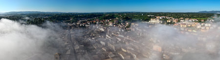 Aerial view of Acquapendente, Italy, shrouded in morning mist. Stone buildings with terracotta roofs are nestled amidst lush greenery under a clear blue sky, highlighting the town's medieval charm.の写真素材