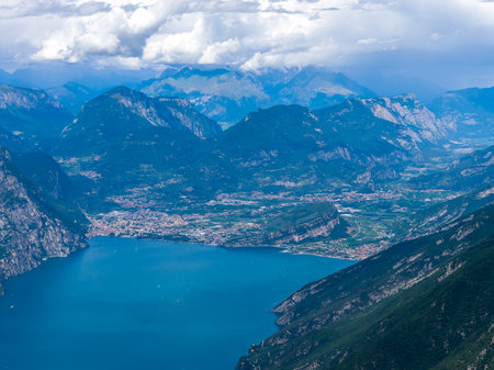 High aerial view of Riva del Garda on Lake Garda, showing alpine slopes, dense lakeside town and cultivated valley under summer clouds.の写真素材