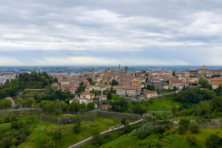 Panoramic view of Bergamo skyline beyond green hills and historic walls under overcast summer sky in Lombardy, northern Italy.の写真素材