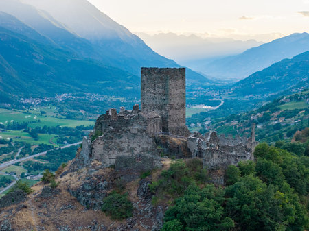 Medieval Castello di Cly stone keep crowns a rocky hill above the Aosta Valley at dusk, overlooking Alpine villages and fields.の写真素材