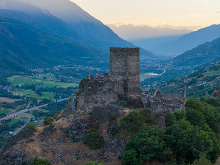 Medieval Castello di Cly tower dominates the Aosta Valley landscape at sunset, stone ruins rising above vineyards and alpine villages.の写真素材