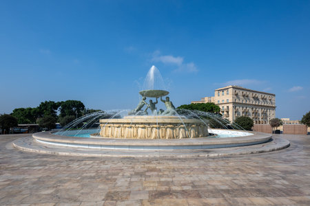 Wide view of the Tritons Fountain in Valletta, Malta, with three bronze tritons holding a basin, water jets, and a clear blue sky in a paved city square.の写真素材