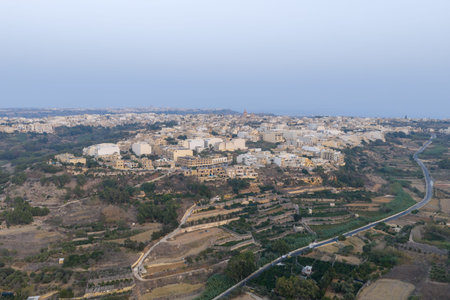 Aerial view of Xaghra town on Gozo, Malta, showing dense limestone buildings on a hilltop with terraced fields, dry valleys, and roads under a clear summer sky.の写真素材