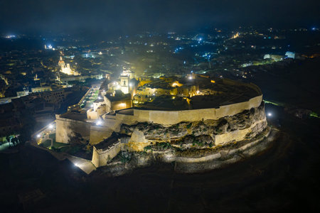 Night aerial view of the Cittadella citadel in Victoria (Rabat), Gozo, Malta, glowing with warm lights against the dark island landscape.の写真素材