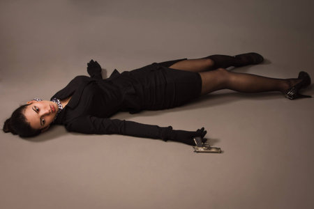 Detective scene imitation. Woman in a black suit with gun lying on the floor