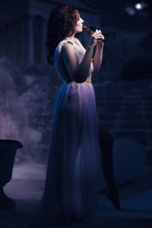 Ancient godness in a white greece toga playing the flute on a temple ruins background. Night lights.の写真素材