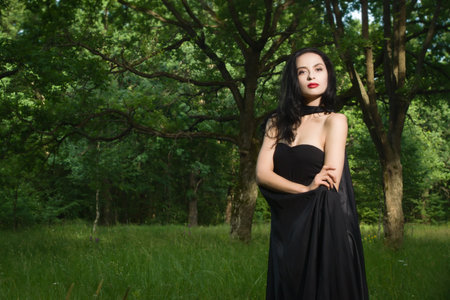 Beautiful brunette woman in black dress and black cloak in the magic forest. Gothic style.の写真素材