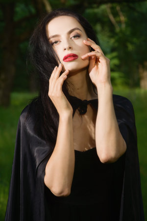 Beautiful brunette woman in black dress and black cloak in the magic forest. Gothic style.