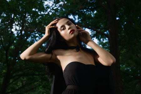 Beautiful brunette woman in black dress and black cloak in the magic forest. Gothic style.の写真素材