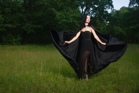 Beautiful brunette woman in black dress and black cloak in the magic forest. Gothic style.の写真素材