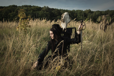 Gothic girl with a sword in her hands next to the skull of a cow on a pole in a summer fieldの写真素材