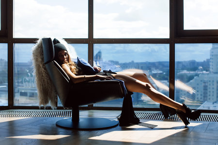 Beautiful girl sitting on a chair her feet up on the window frame in a lace bodysuit, a blue coat and wearing shoes with heels can be seen outside the city. On a chair thrown over the white skinの写真素材