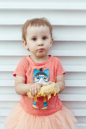 A little girl eaten corn on the cob soiled, grubby on a light background in a pink shirt and tutu skirtの写真素材