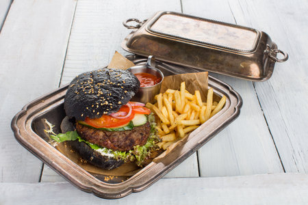 Black burger with fries and ketchup, served in a metal container with a patina on white wood tableの写真素材