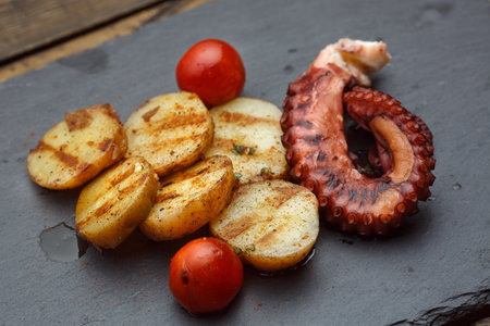 Tasty grilled potatoes with seafood octopus and tomato on a slate plate on a wooden tableの写真素材