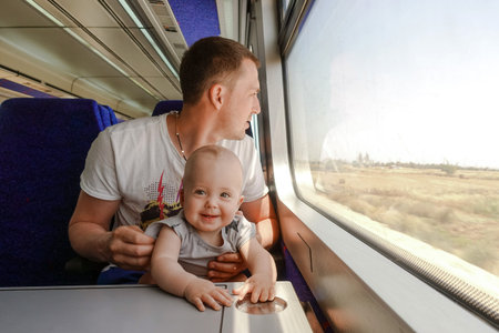 Young man - father, travelling with his little son by train, looking at the window. Dad with a laughing baby sitting at the table in the interior of the trainの写真素材