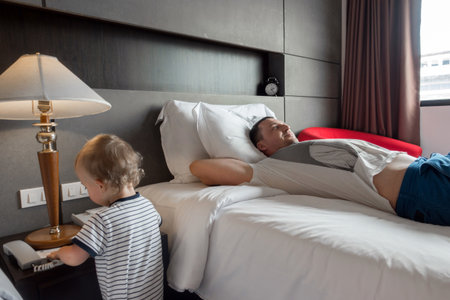 Content father lying on cozy bed in hotel room with cute infant playing near.の写真素材