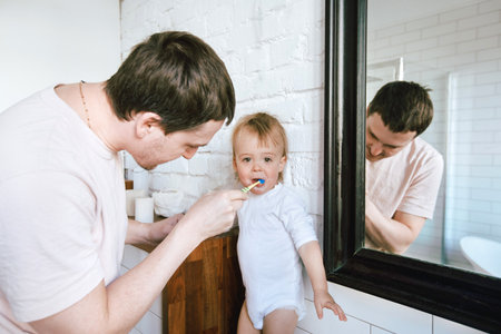 Happy father helping with teeth brushing to charming little boy in light white bathroom.の写真素材