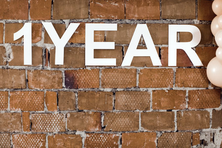 Arranged white letters saying One year on shabby brick wall with balloons near.