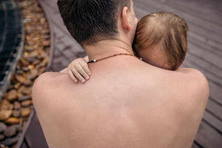 Back view of shirtless man embracing little child sitting on poolside after swimming.の写真素材