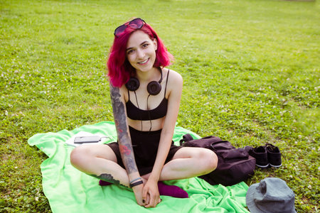 Young informal woman with pink hair and tattoos sitting on plaid in green park squinting eyes from happiness looking at cameraの写真素材