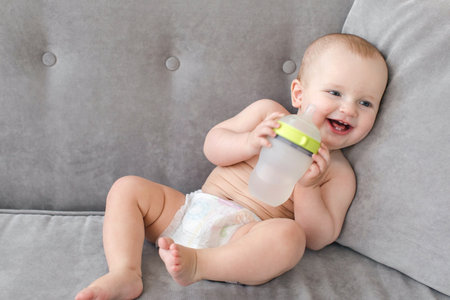 Cute baby with bottle of water lying on comfortable couch and laughing