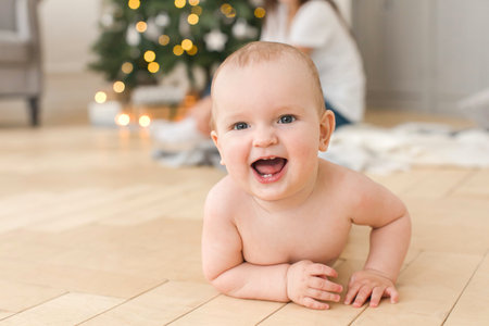 Smiling naked baby lying on floor and looking at camera on blurred background of Christmas tree in...