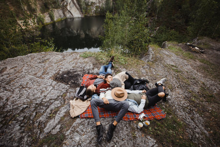 From above view of group of friends lying on plaided blanket on ground near steep leading to lakeの写真素材