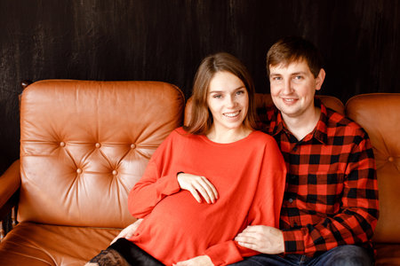 Handsome man and lovely pregnant woman smiling and looking at camera while sitting on comfortable couch togetherの写真素材