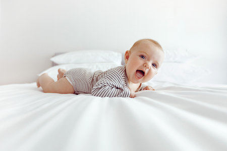 Side view of adorable little baby lying on white soft blanket on bed smiling excitedly in daylight
