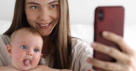 Beautiful young woman smiling and taking selfie with funny baby while spending time at home